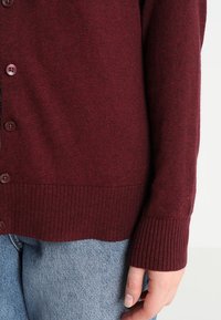 Cardigan en maille bordeaux avec poignets et ourlet côtelés, doté de boutons ronds. Le tissu présente une finition légèrement texturée.