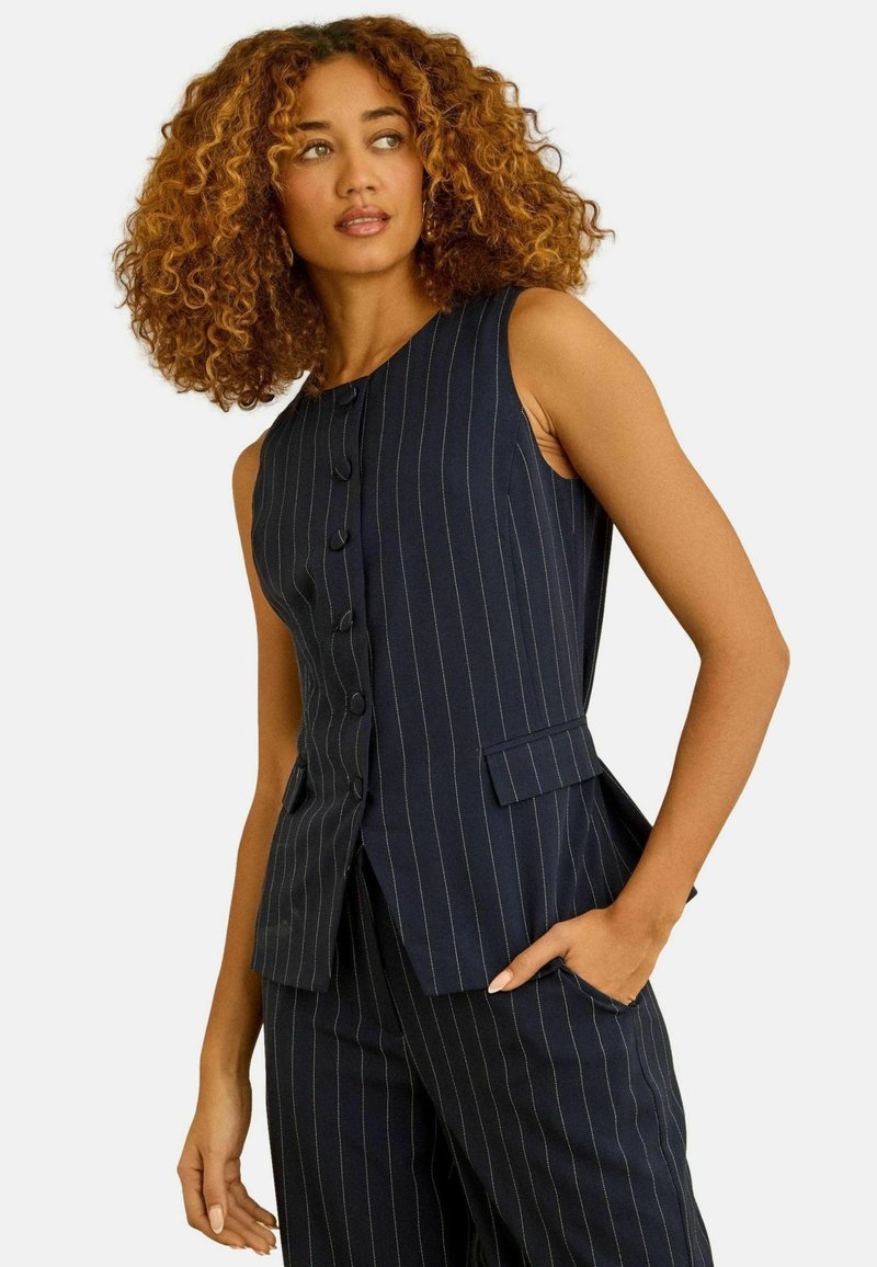 Woman with curly hair wearing navy blue pinstripe sleeveless buttoned vest and matching trousers, hand in pocket, looking to the side.