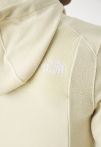 Sudadera de forro polar beige con una superficie texturizada. Presenta un logo bordado blanco en el hombro izquierdo y un bolsillo tipo canguro.