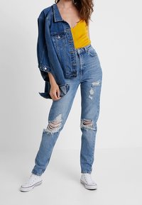 Jeans de denim azul claro con detalles desgastados y una cintura alta, combinados con una blusa amarilla y una chaqueta de denim. Zapatillas blancas completan el look.