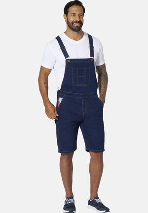 Man smiling wearing white t-shirt, dark blue denim short overalls, and navy sneakers, hands in pockets on light background.