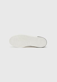 Sola de sapatilha branca com padrão de aderência texturizado, apresentando design em espinha de peixe, e contorno preto no calcanhar para destaque.