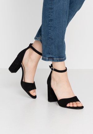 Sandales à talons hauts en suède noir avec un design à bout ouvert, une bride de cheville avec une boucle en acier inoxydable et un talon carré large.