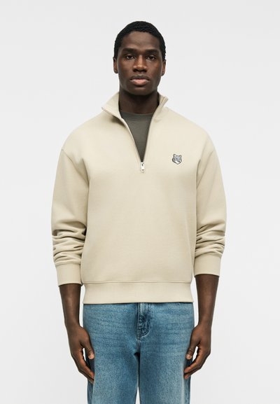 Jeune homme portant un sweat-shirt beige à quart de fermeture éclair avec un logo de loup sur la poitrine et un jean bleu, debout devant un fond uni.