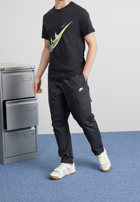 Černé tričko Nike se zeleno-bílým logem, doplněné černými cargo kalhotami a bílými teniskami se žlutými tkaničkami a gumovou podrážkou.