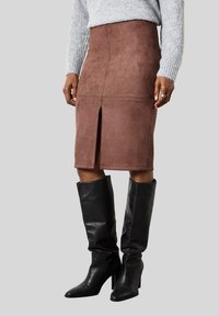 Femme portant une jupe en daim marron jusqu'aux genoux avec une fente à l'avant, un pull en tricot gris et des bottes à talons noires montant jusqu'aux genoux sur fond gris.