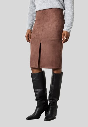 Femme portant une jupe en daim marron jusqu'aux genoux avec une fente à l'avant, un pull en tricot gris et des bottes à talons noires montant jusqu'aux genoux sur fond gris.