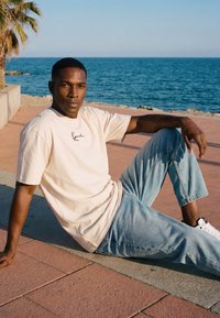 Mies beige t-paita ja vaaleansiniset farkut päällä istuu päällystetyllä polulla meren lähellä, taustalla on palmupuu.