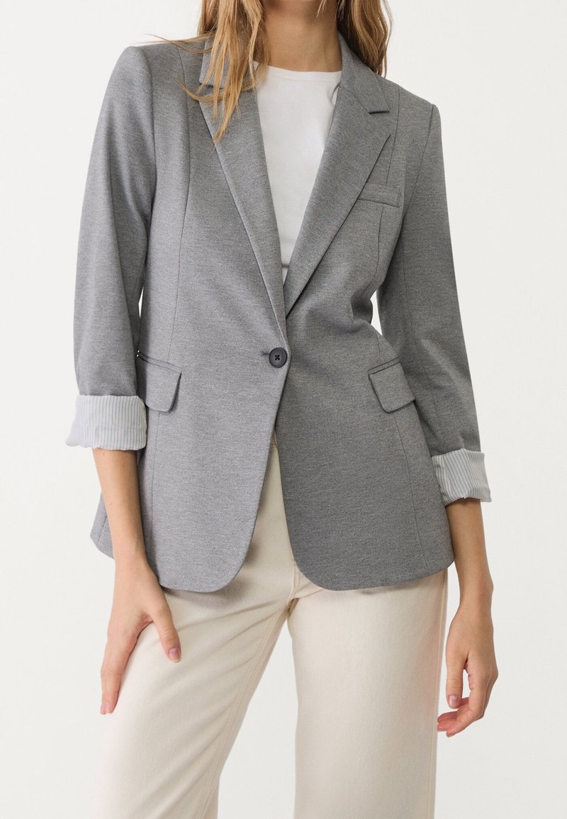 Femme portant un blazer gris à un bouton avec les manches retroussées sur une chemise blanche et un pantalon beige clair sur un fond uni.