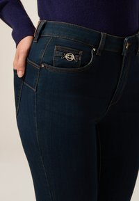Jeans de mezclilla oscura con un diseño ajustado, que presentan un detalle decorativo en el bolsillo con un adorno metálico y costuras en contraste.