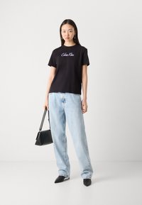 Crna pamučna majica s "Calvin Klein" logotipom, svijetloplave traperice, crna torba i crne cipele s oštrim vrhom. Minimalistički dizajn.