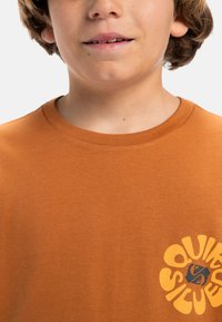 Oranje katoenen T-shirt met een ronde halslijn en korte mouwen, met een gele cirkelvormige grafiek met zwarte accenten op de rechteronderkant.