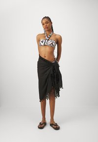 Zwarte sarong met franjes, gecombineerd met een wit en zwart gestreept bikinitopje met halternek en groene accenten.