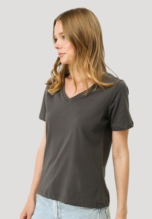 Frau mit blonden Haaren, die ein enges, dunkelgraues V-Ausschnitt-T-Shirt und hellblaue Jeans trägt, schaut nach links vor einem schlichten Hintergrund.