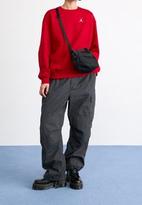 Person trägt ein rotes Sweatshirt, schwarze Cargohosen, schwarze Plateaustiefel und eine schwarze Umhängetasche, steht auf einem blauen Boden.