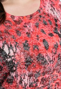 Camisa de patrones rojos y rosas con un escote redondo, que presenta diseños abstractos en negro y gris, además de adornos metálicos. Textura de tela suave.