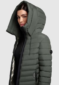 Grüne Steppjacke mit großer Kapuze, Reißverschluss vorne und seitlichen Taschen. Verfügt über eine gesteppten Struktur und schwarze Metallakzente.