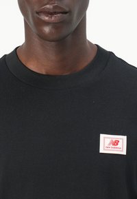 T-shirt en coton noir avec un col rond. Il présente une petite étiquette rectangulaire blanche avec le logo rouge "New Balance" sur la poitrine.