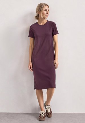 Femme aux cheveux blonds en chignon bas portant une robe midi violette à manches courtes et des baskets marron, debout devant un mur clair uni.
