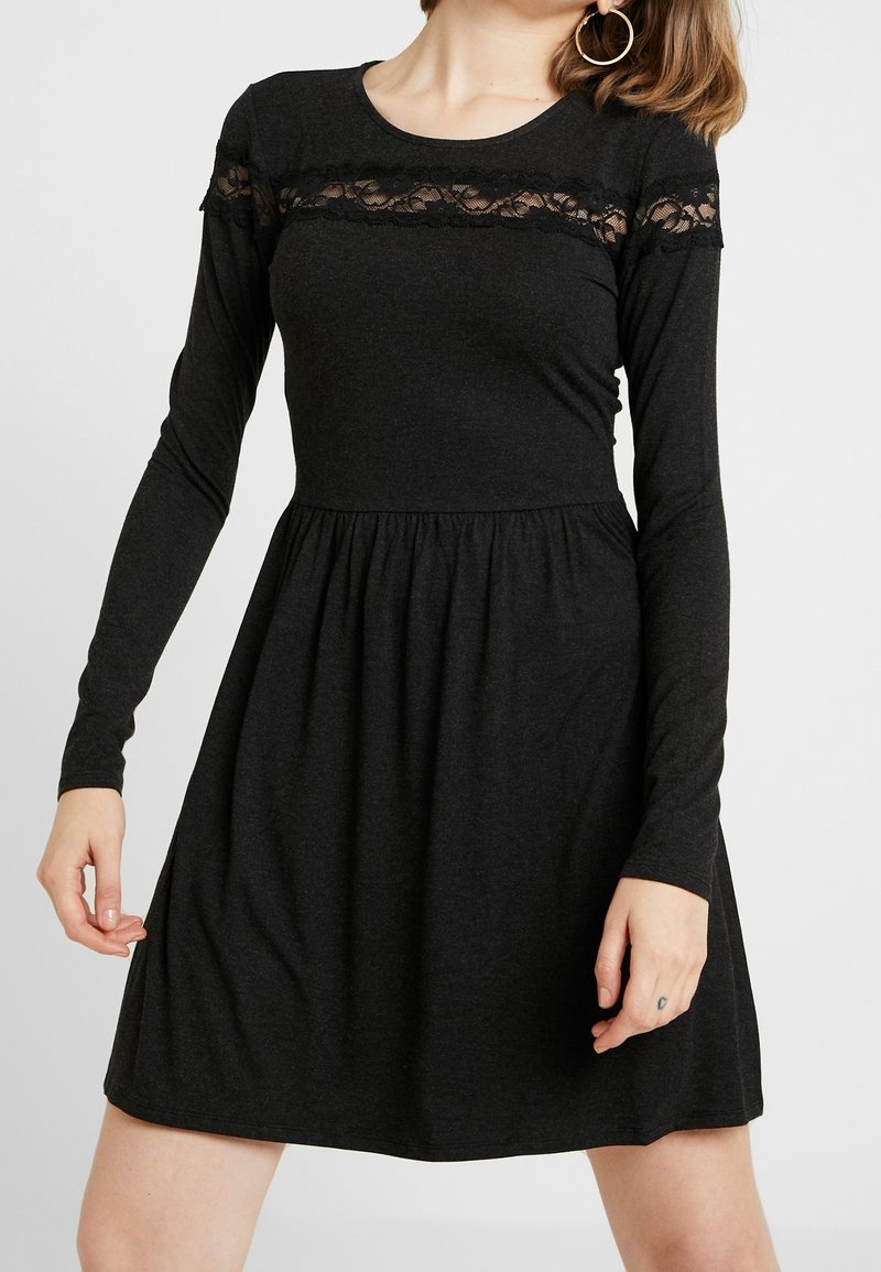 Femme portant une robe noire à manches longues avec un détail en dentelle sur la poitrine et une jupe froncée, debout devant un fond uni.