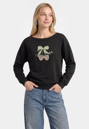Giovane donna che indossa una felpa nera con grafica di fiocco lucido e ciliegie, orecchini a cerchio blu e jeans blu chiaro con le mani nelle tasche.