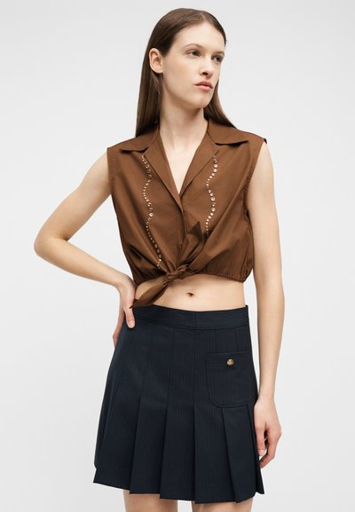 Femme aux cheveux bruns raides portant un haut court marron sans manches noué à l'avant et une jupe plissée noire avec poche avant, posant avec la main sur la hanche.