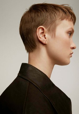 Cabello corto y castaño claro, perfil lateral; lleva una chaqueta oscura y Texturizada con un cuello pronunciado. Se ve un pequeño aro de oro en la oreja.
