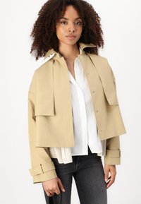 Veste beige cropped avec un col surdimensionné, des fermetures à pression et de grandes poches avant. Tissu texturé, manches larges et poignets ajustables.