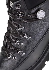 Bota de senderismo de cuero negro con superficie texturizada, ojales de metal y cordones grises. Suela de goma con detalles de relieve. Acento de logo en el costado.