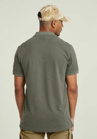 Polo shirt verde scuro con maniche corte, realizzato in tessuto misto cotone. Presenta un colletto classico e un orlo dritto. Cappellino beige a motivo camouflage indossato.