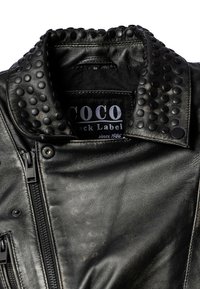 Schwarze Lederjacke mit Nietenkragen, Reißverschluss und strukturiertem Oberflächen. Das Etikett trägt die Aufschrift "COCO Black Label seit 1986."