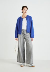 Woman wearing blue buttoned cardigan, white ribbed top, wide gray jeans, silver sneakers, and holding a white handbag against white background.