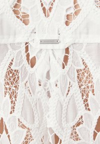 Balts mežģīņu apģērbs ar sarežģītiem ziedu un ģeometriskiem rakstiem un izgriezumiem; tajā iekļauta maza metāla zīmīte ar uzrakstu "ALLEGANTE".