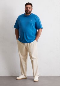 Blauwe katoenen t-shirt met korte mouwen, gecombineerd met beige relaxed-fit broek en witte sneakers. Eenvoudig ontwerp, zonder zichtbare patronen of graphics.