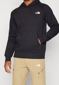 Sudadera negra con capucha, bolsillo frontal y puños acanalados, con un logo blanco en el lado izquierdo del pecho. Combinada con pantalones beiges.