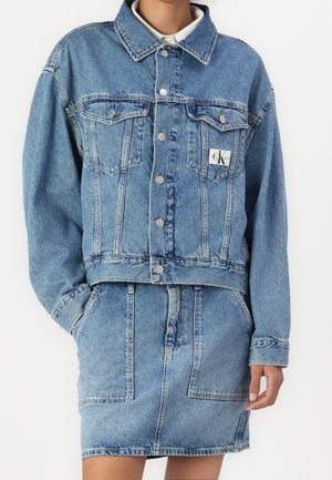 Kvinde iført lyseblå denimjakke med sølvknapper og matchende denimnederdel med forlommer, hænderne i lommerne.