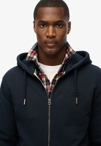 Hoodie bleu marine à fermeture éclair avec capuche à cordon, superposé sur une chemise en flanelle écossaise rouge et verte. Logo sur la poitrine et détail de la fermeture éclair argentée.