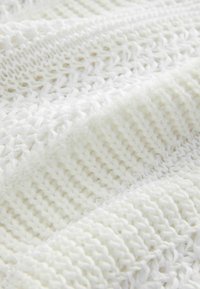 Vitt stickat tyg med varierande texturer och mönster, inklusive täta vävningar och lösare stygn, som visar en mjuk och fluffig känsla.