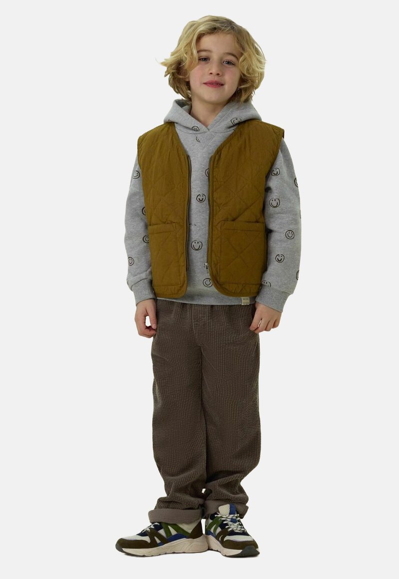 Braune gesteppte Weste über einem grauen Hoodie mit Smileymuster, kombiniert mit braunen Cordhosen und mehrfarbigen Sneakers.