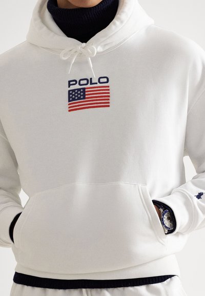 White pullover hoodie with front pocket, featuring "POLO" text and U.S. flag design. Fabric is smooth with a soft texture. Ribbed cuffs.