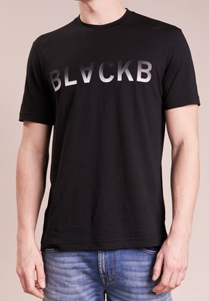 Schwarzes kurzärmeliges T-Shirt mit silbernem Farbverlauftext "BLACKB", getragen zu blauen Jeans, zeigt den oberen Torso und die Arme.
