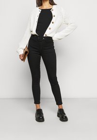 Gilet en tricot blanc avec des boutons, haut ajusté noir, jean noir taille haute et chaussures noires à lacets. Tient une pochette marron.
