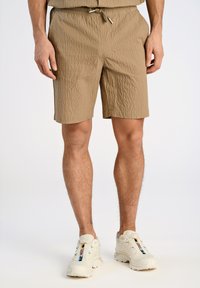 Pantalones cortos beige, de longitud hasta la rodilla, ligeros y con una superficie texturizada, cinturilla elástica y cordón. Llevados con zapatillas deportivas blancas.