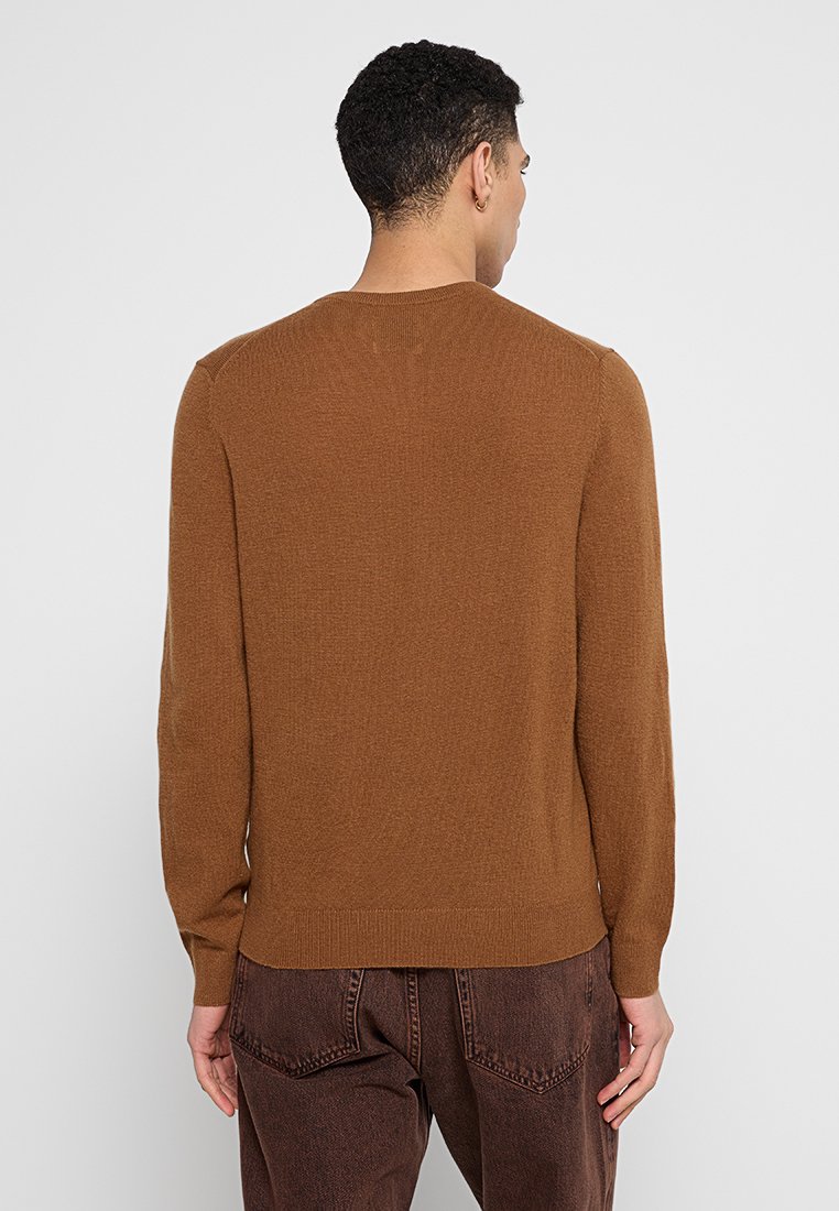 Mann mit kurz lockigem Haar, der einen braunen Pullover mit langen Ärmeln und dunkelbraune Jeans trägt, und mit dem Rücken zu einem schlichten hellen Hintergrund steht.