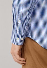 Mannelijke hand rustend naast de blauw-witte geblokte manchet van een shirt met twee knopen, gecombineerd met een bruine broek.