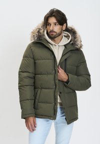 Oliven grøn polstret jakke med en pelsforet hætte, der har en lynlås og trykknaplukning, samt en brystlomme med lynlås.