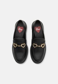 Mustad musta nahast loaferid, millel on kuldne südameketi aksessuaar, sujuva tekstuuri ja ümar disainiga. Sees on punane südame logo tallal.