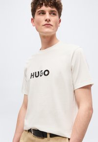 Νεαρός άνδρας με σγουρά μαλλιά φοράει λευκό μπλουζάκι με το λογότυπο HUGO, μπαίνει μέσα σε μπεζ παντελόνια με μαύρη ζώνη, κοιτάζει ελαφρώς προς τα δεξιά.