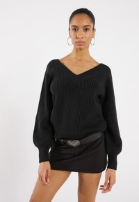 Schwarzer, gerippter Strickpullover mit V-Ausschnitt und voluminösen Ärmeln, getragen über einem schwarzen Minirock. Hat eine silberne, herzförmige Schnalle.