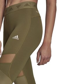 Olijfgroene aansluitende leggings met een hoge tailleband, details van netstof en een wit Adidas-logo aan de linkerkant. Zachte, rekbare stof.